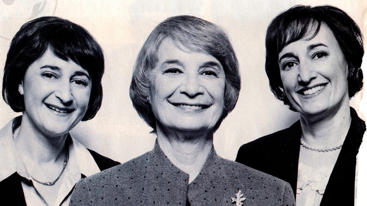 A mother stands flanked by two daughters and they smile at the camera.