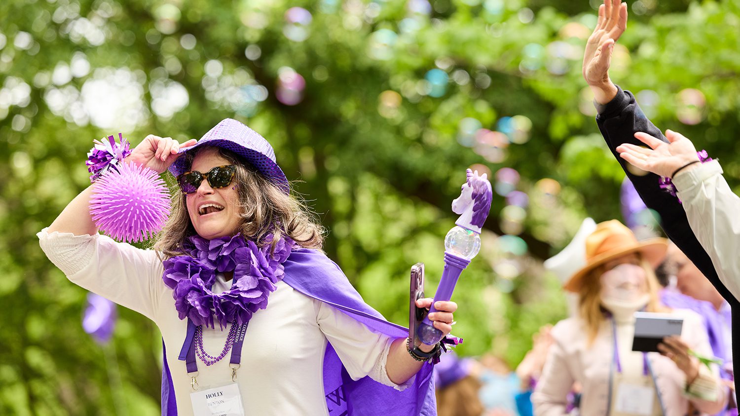 A member of the class of 1990 in a purple hat smiles amid a cloud of bubbles