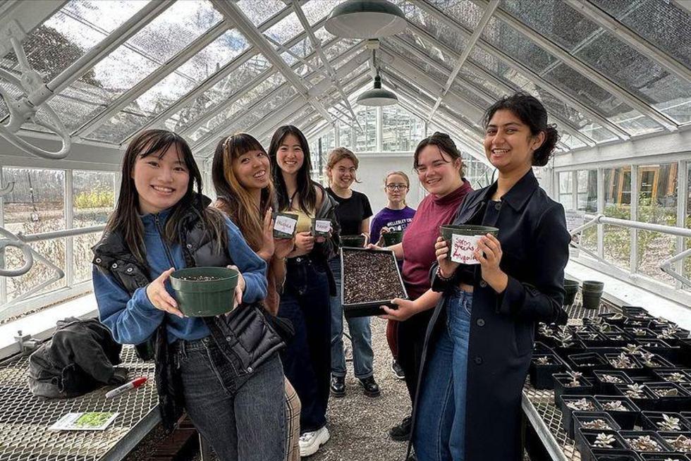 Seven students standing in a greenhouse smile. They are holding pots with dirt.