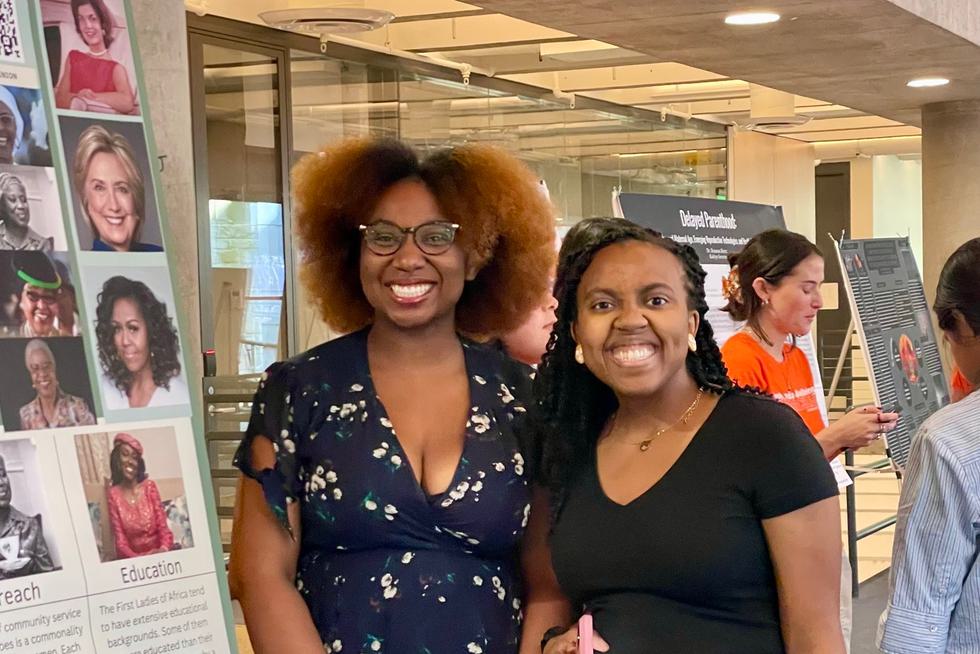 Two students stand next to a research poster with images of Hillary Clinton and Michelle Obama on it. Both students are smiling.