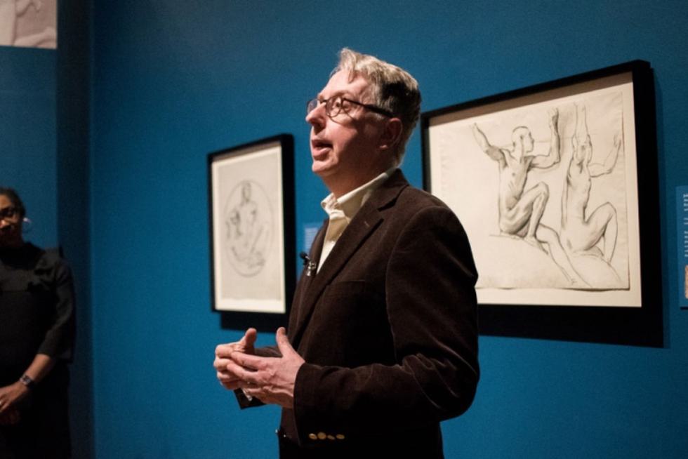 Professor Paul Fisher stands and talks in a museum in front of two drawings on a blue wall.