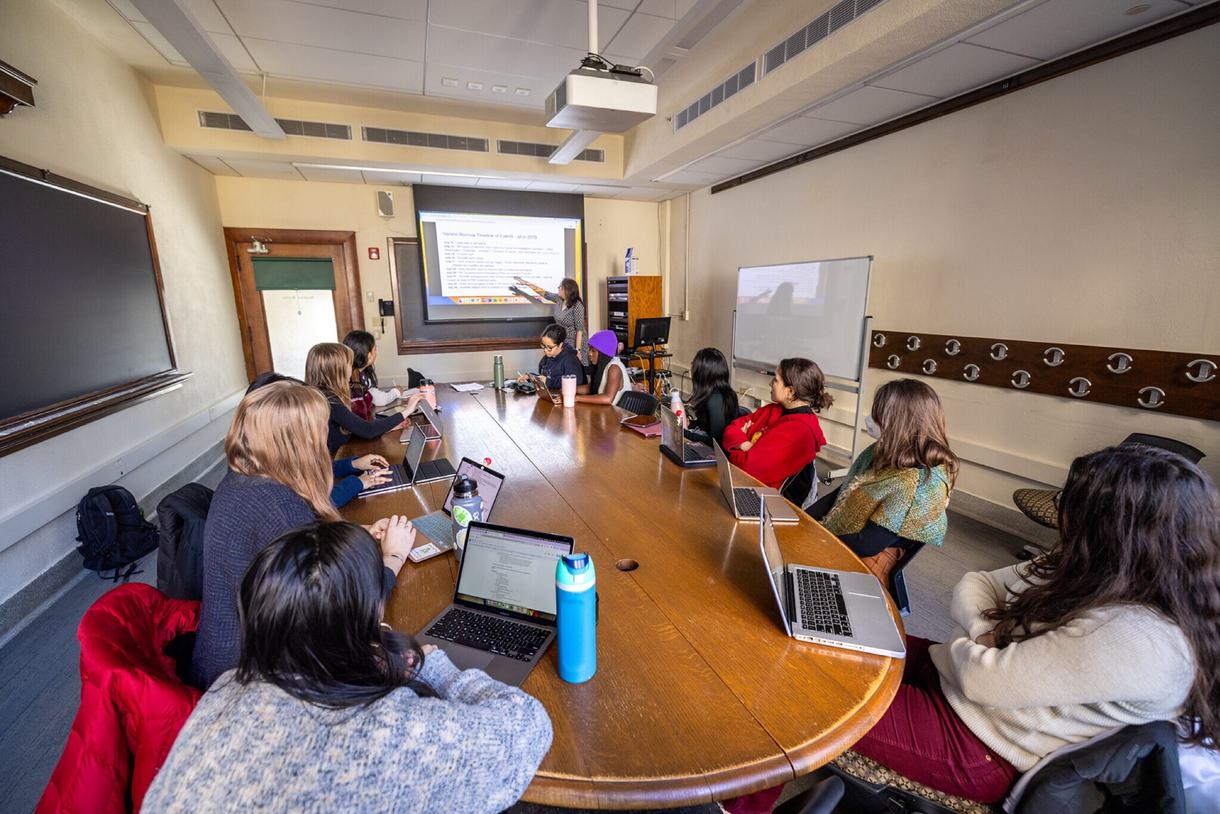 Eleven students sit around a table in a classroom and watch as the professor points to the screen with bullet points on it.