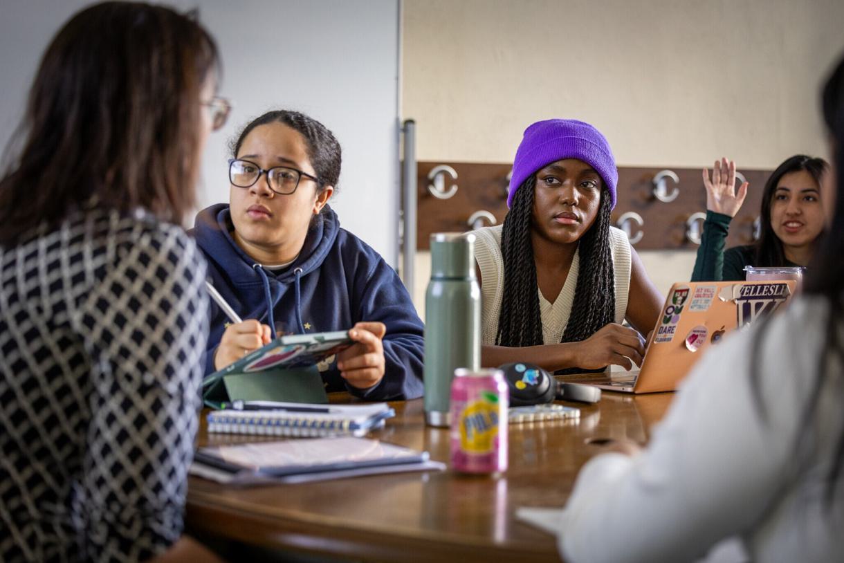 Two students sitting at a classroom table watch intently as a professor talks to them with her back turned. There is a third student raising her hand in the background.