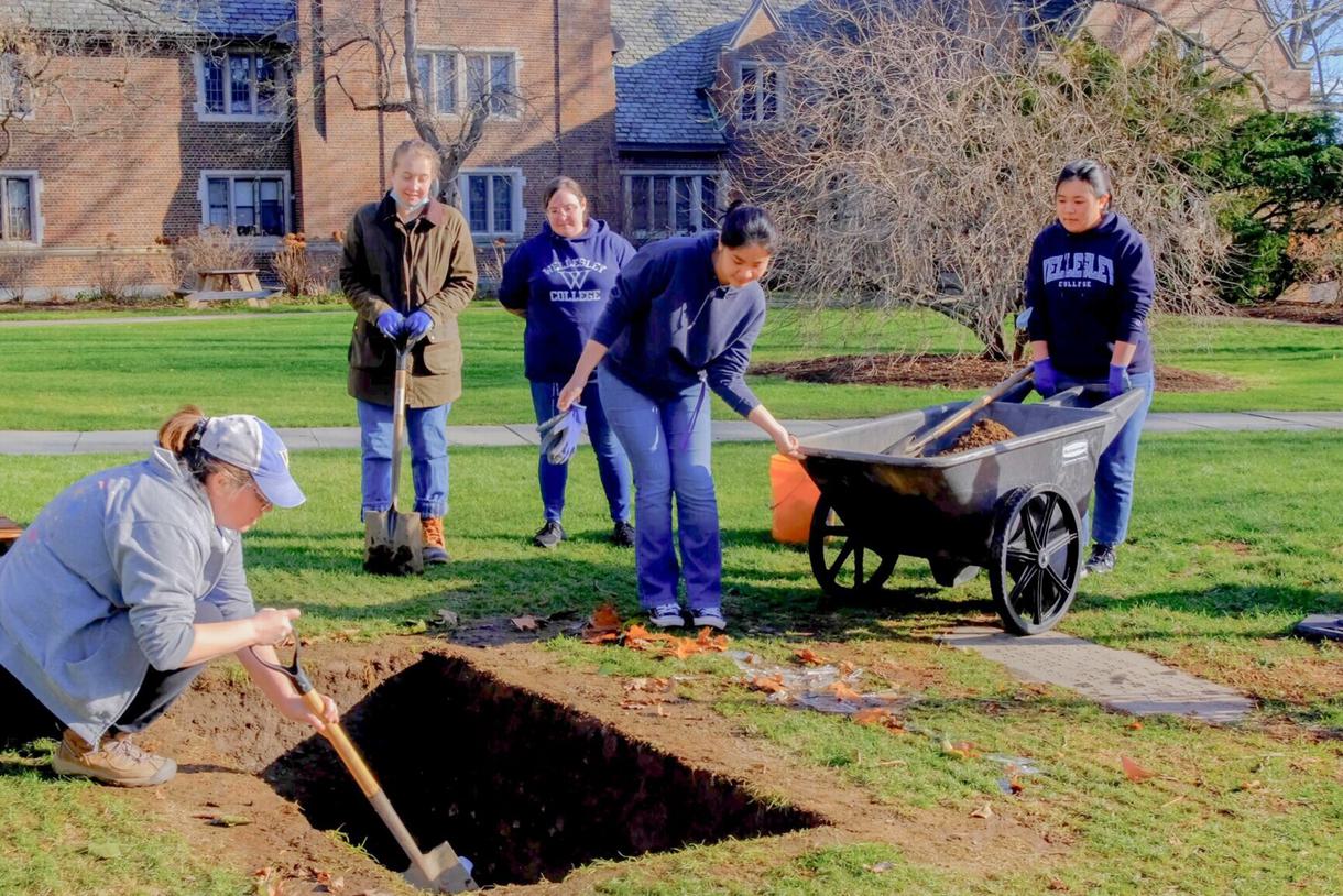 4 students are around a hole in the ground that is being dug by a fifth student. They are wearing work gloves and jeans.