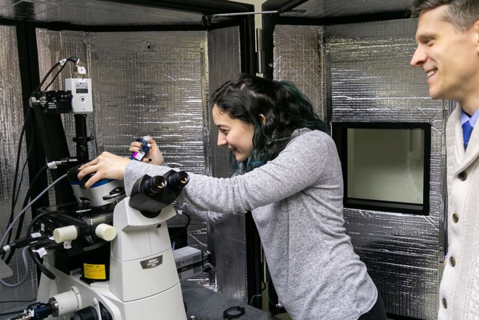A student works on a microscope while a professor observes. Both are smiling.