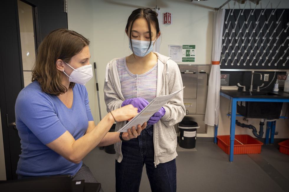 A professor holds a piece of paper that they are showing a student, who is standing to her left. Both individuals are wearing masks, and the student is wearing purple gloves.