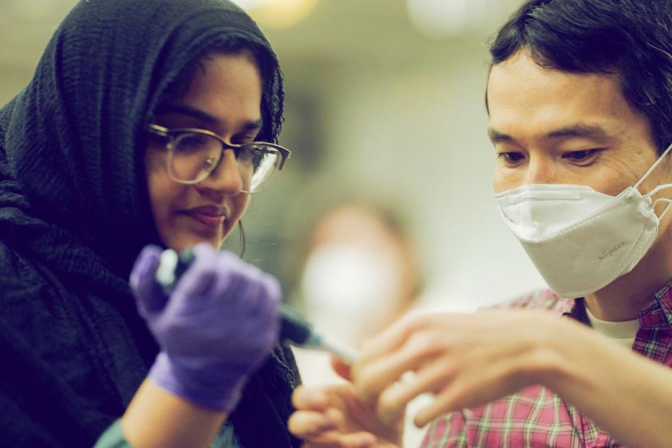A student wearing a hijab helps a professor wearing a mask with a titration lab.