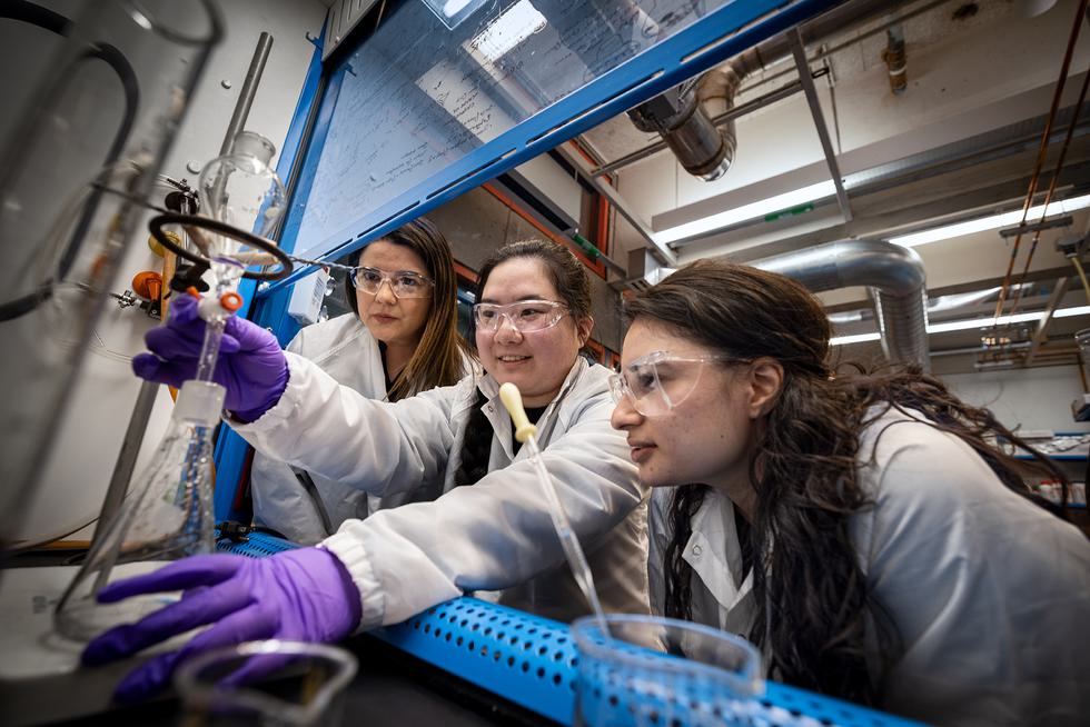 Three students dressed in lab gear lean into a fume hood and work on a titration.