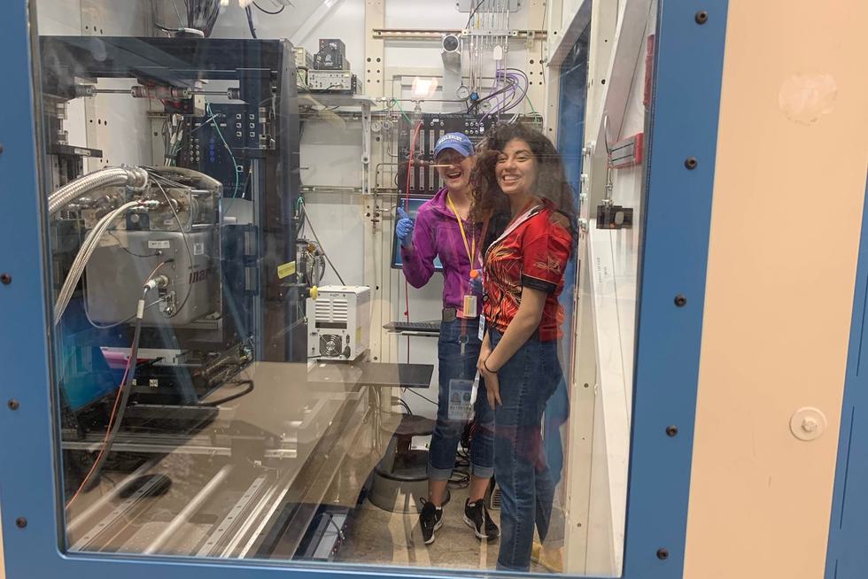 Two students work at a 3D printer. One of them is holding a thumbs-up.