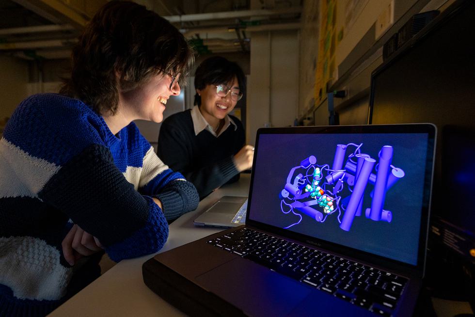 Two students in a dimly lit room look at an open laptop. Another open laptop shows a 3-D model of a molecule.