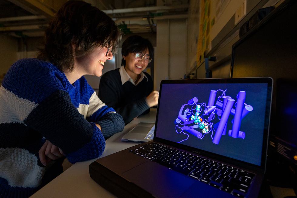 Two students smile while looking at a laptop. Another laptop screen shows a rendering of a peptide.