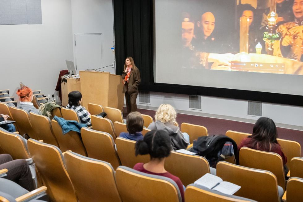A professor lectures at the front of an auditorium with a still from a film projected on a screen behind her