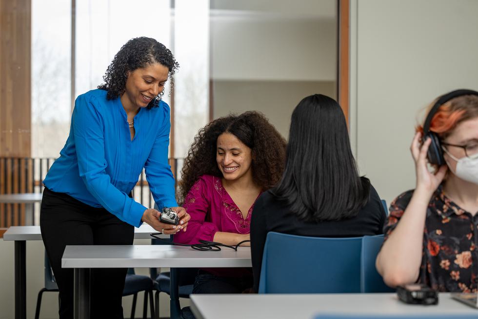 Professor Sabriya Fisher shows a student how to operate a recording device. The professor is standing and the student is sitting. Both are smiling.