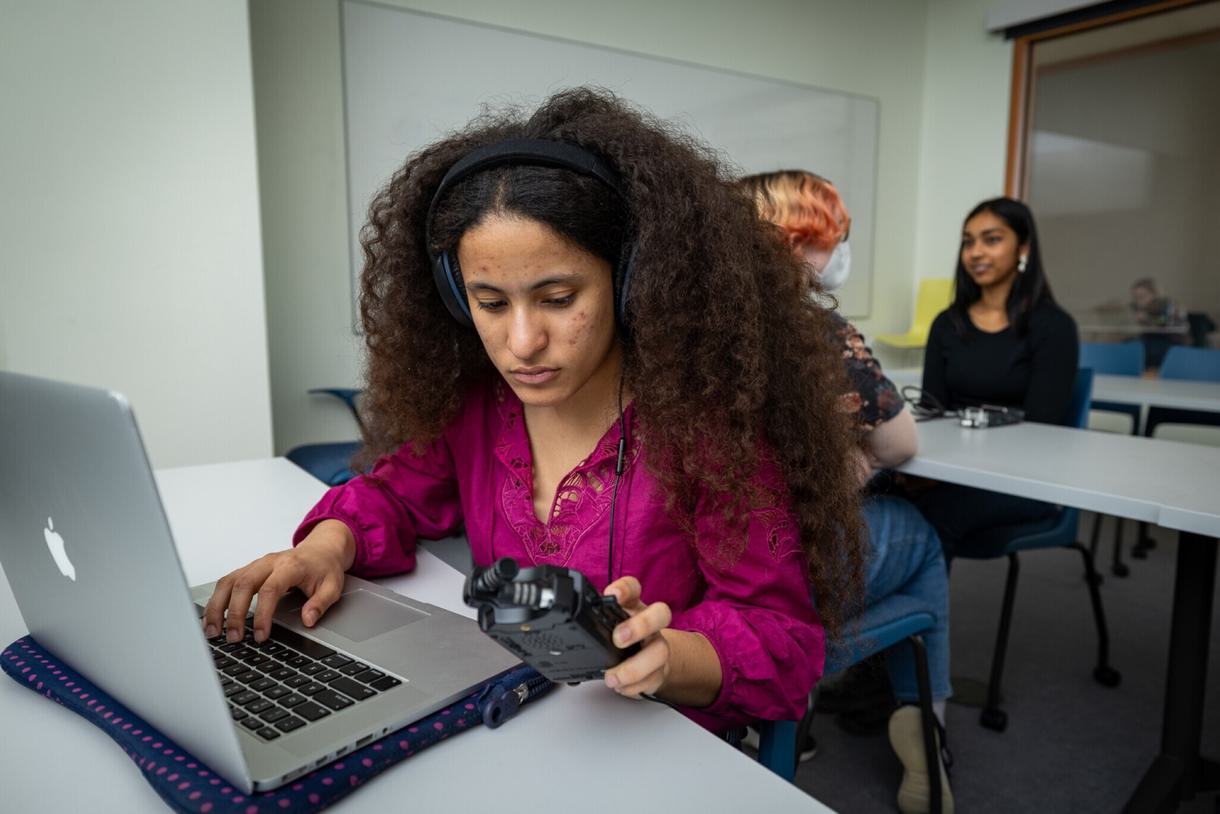 A student wearing a pink shirt and headphones listens to a recording device and transcribes on her laptop.
