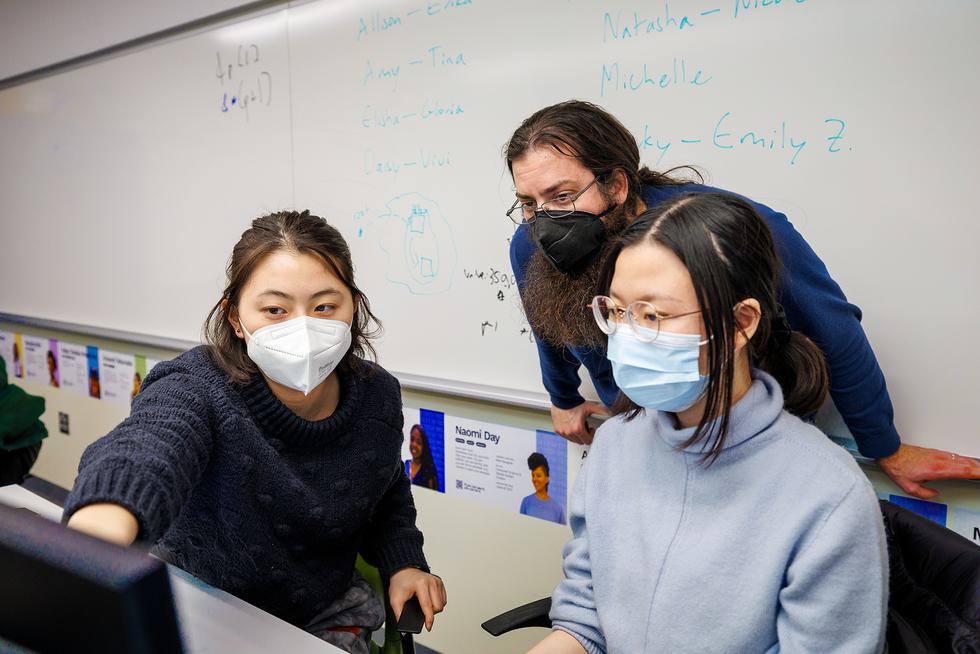 Two students wearing masks sit at a computer desk. One student is pointing to the screen of a computer while a professor leans over them, also wearing a mask.