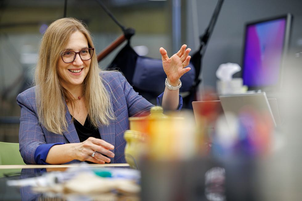Orit Shaer sits at a desk and gesticulates while smiling. She is wearing a purple blazer.