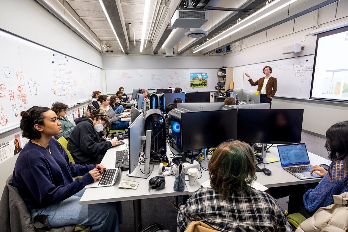 A professor stands at the front of a full computer lab and speaks to students while they are working on desktop computers.