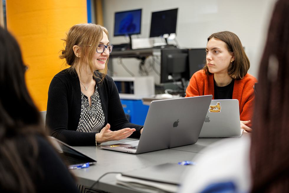 Professor Eni Mustafaraj sits at her laptop while talking to students at a table.