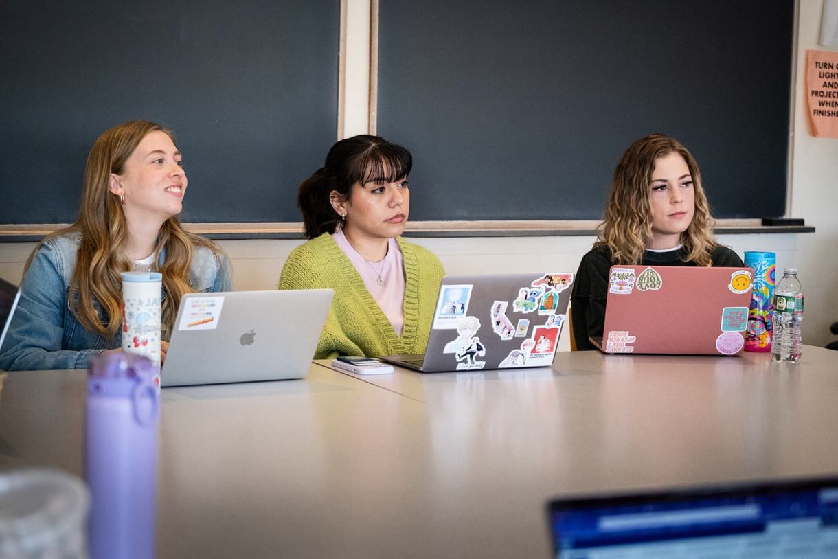 Three students sit in front of a blackboard on their laptops.