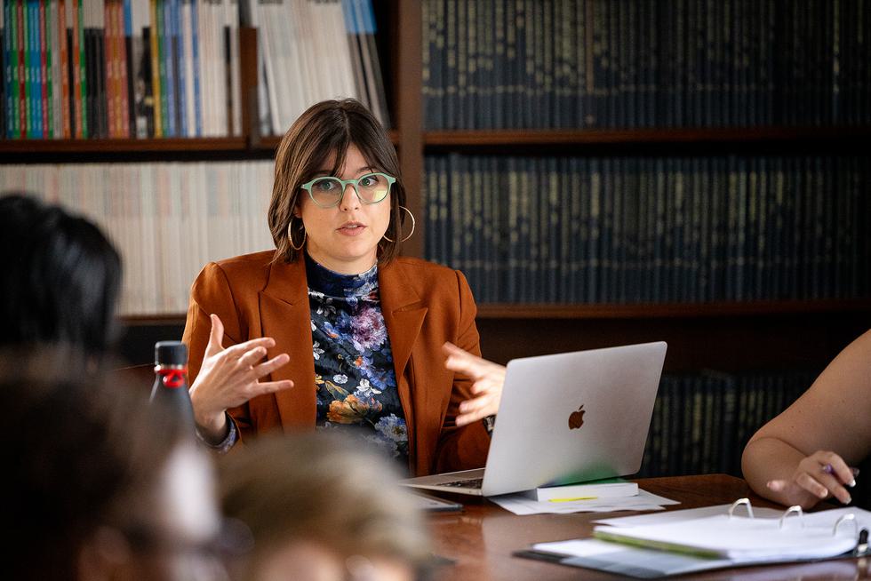 Pamela D’Andrea Martínez sits at a table in front of a bookshelf and speaks to students. She is sitting in front of a laptop, wearing an orange blazer and green glasses.