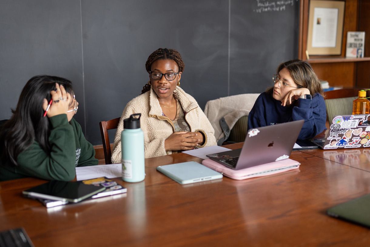 Three students sit at a table in front of their laptops and collaborate on a group project.