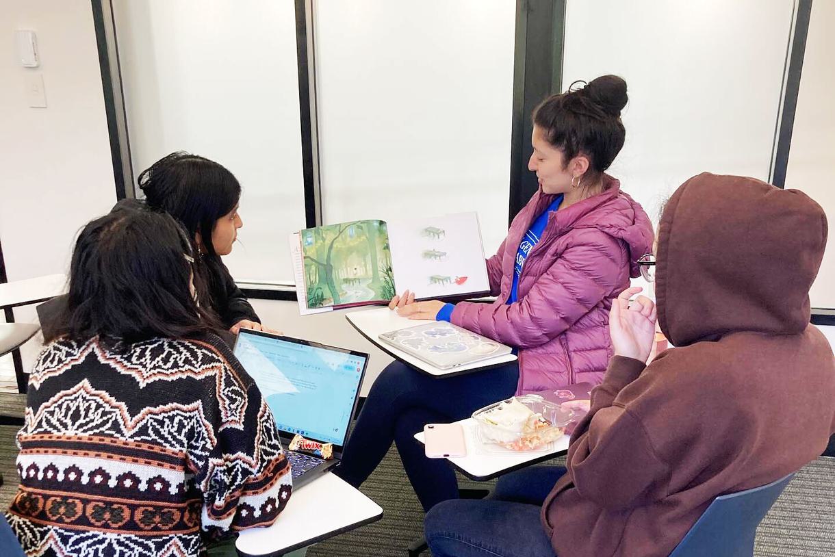 Students sit in a group at movable desks while one reads from a picture book out loud.