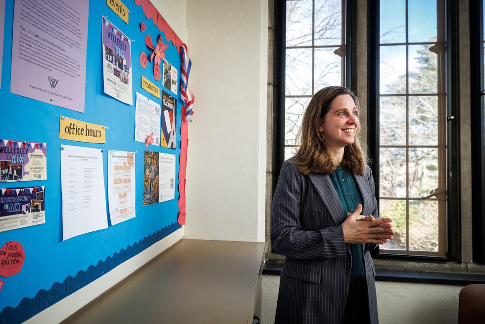 Hélène Bilis stands in front of a colorful bulletin board and lectures to students offscreen. She is smiling and wearing a pinstripe blazer.