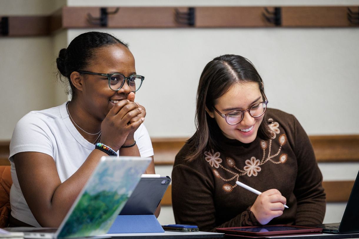 Two students sit in front of iPads taking notes in class. Both are smiling and looking at something off camera.