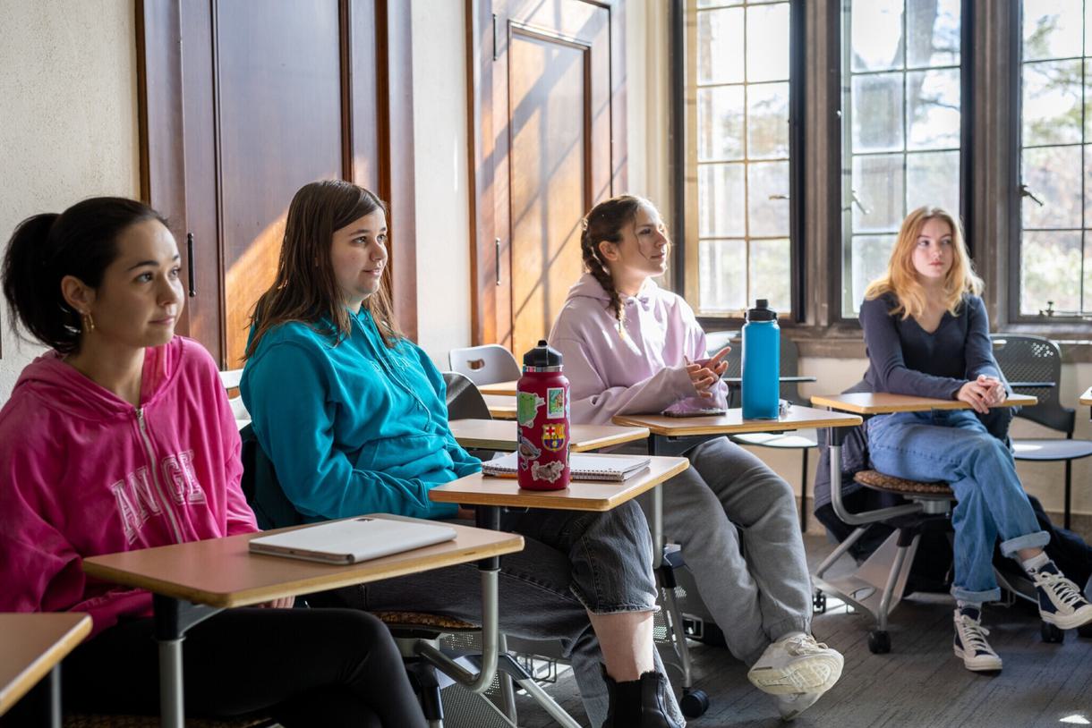 Four students sit at desks in a classroom. They are watching something off screen.