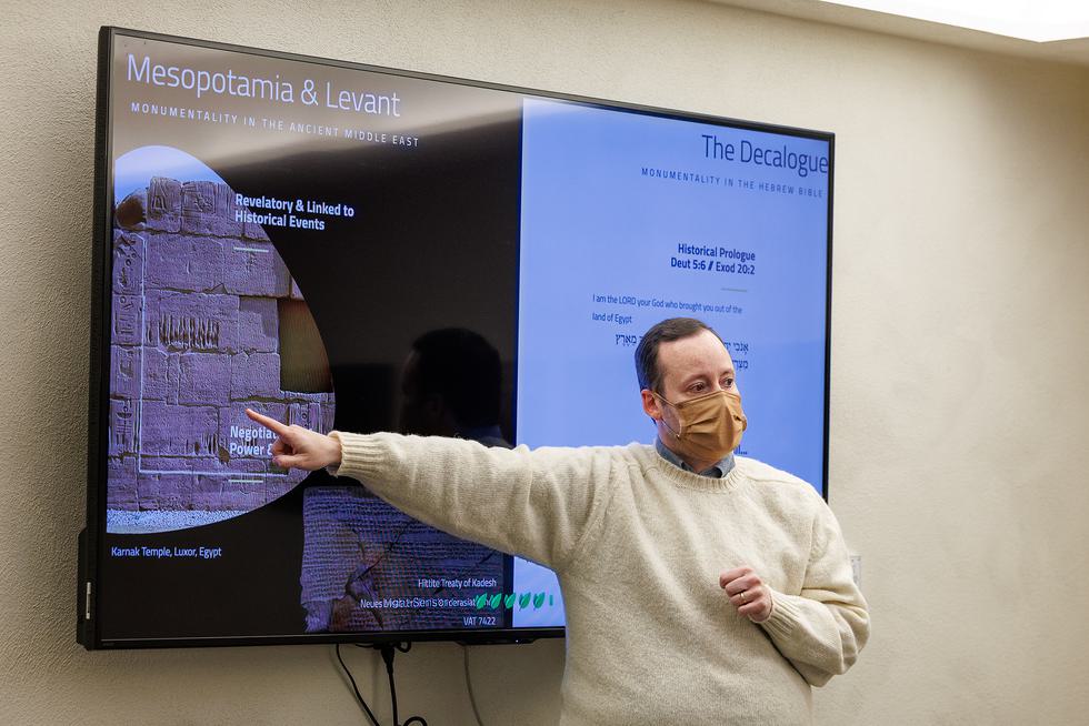 Eric Jarrard, wearing a mask, points at a TV screen with a slide about Mesopatamia and Levant.