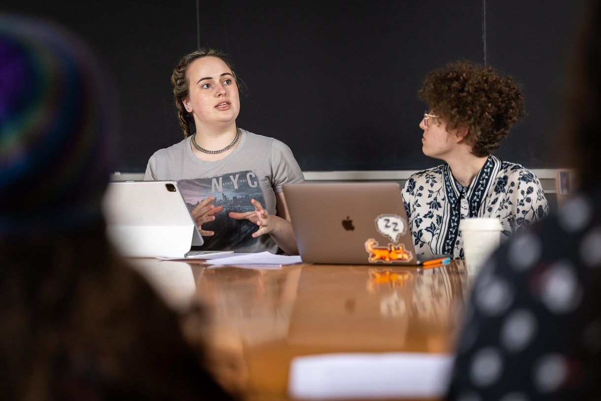 One student speaking and gesturing, another listening, seated with others at a table