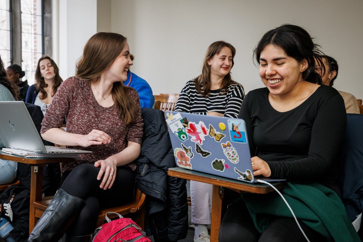 Two students, one with a laptop covered in stickers, laugh with eachother in a classroom. There are other students in the background working on their own laptops.
