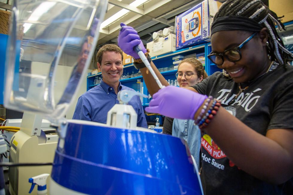 A student pipettes into a container while another student and professor smile behind them.