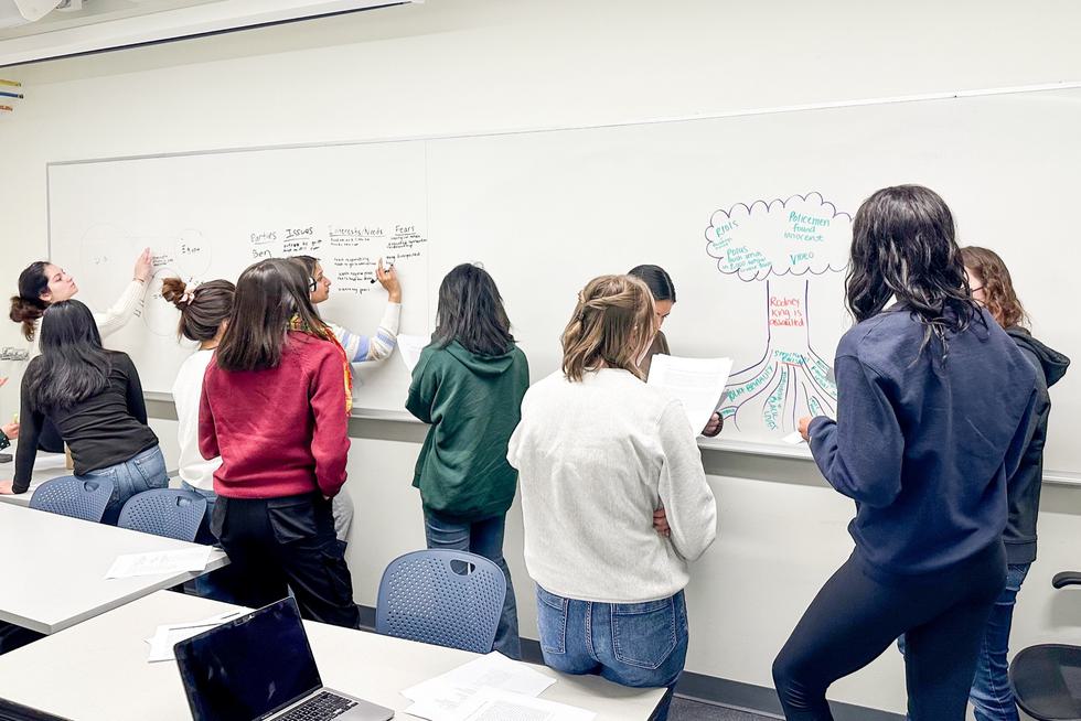 Students in a classroom talk in groups and write on a white board.