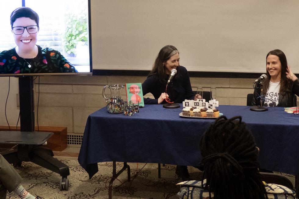 Three presenters, one on a monitor and two at a table, present on a Professor's recently published book.