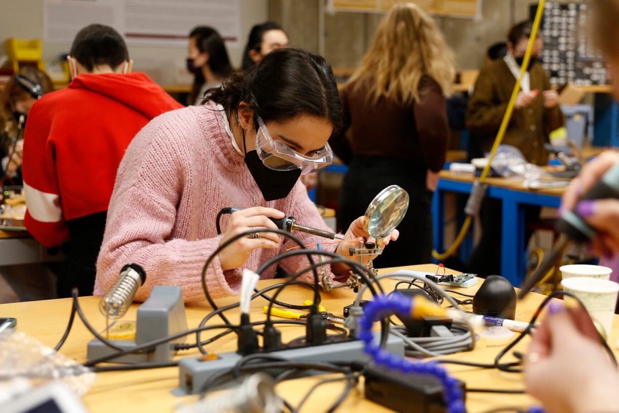 Students wearing protective glasses work at soldering stations in a classroom.