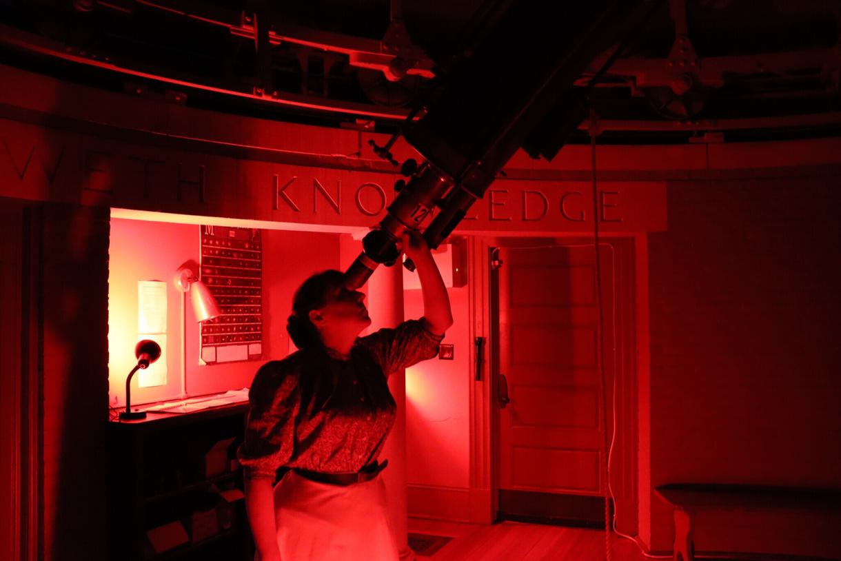 A student uses through the telescope inside Whitin Observatory. The room is drenched in red light.