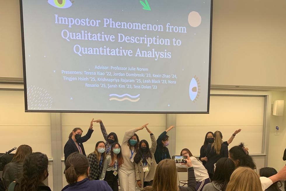 Students pose for a picture in front of a slide reading "Imposter Phenomenon from Qualitative Description to Quantitative Analysis"