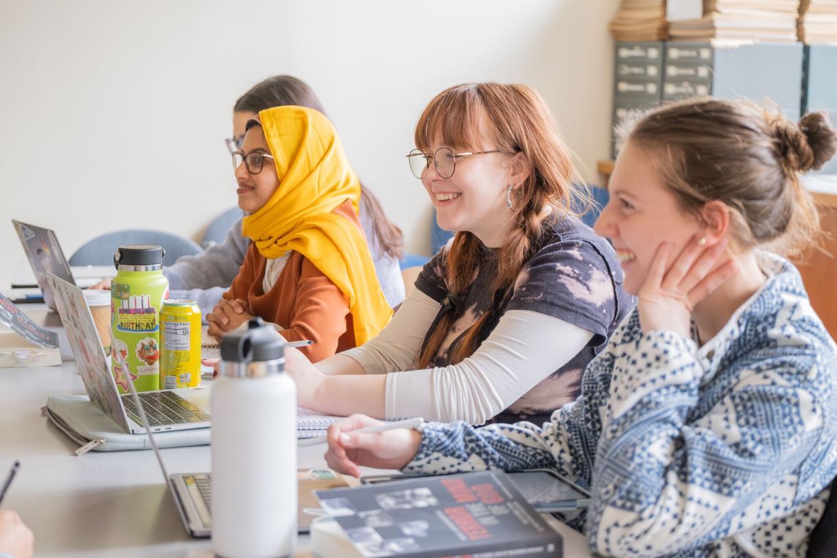 Four students, one wearing a bright yellow hijab, smile and sit on their laptops.