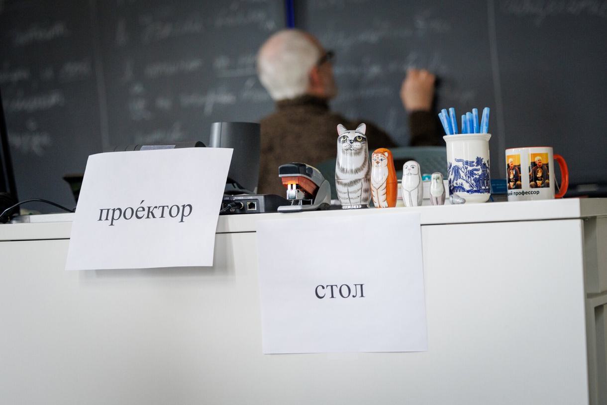Closeup of Russian words taped to a desk in a Russian classroom. A professor writes on the chalkboard behind the desk.