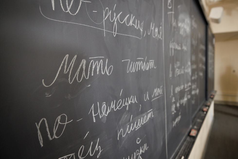 Closeup of Russian words written on a chalkboard. A professor stands off to the side of the chalkboard.