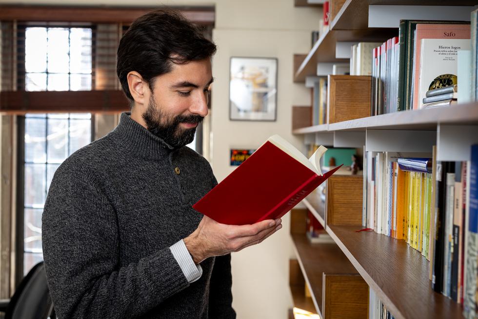 Antonio Arraiza-Rivera reads a book while standing at a bookshelf. He is softly smiling.