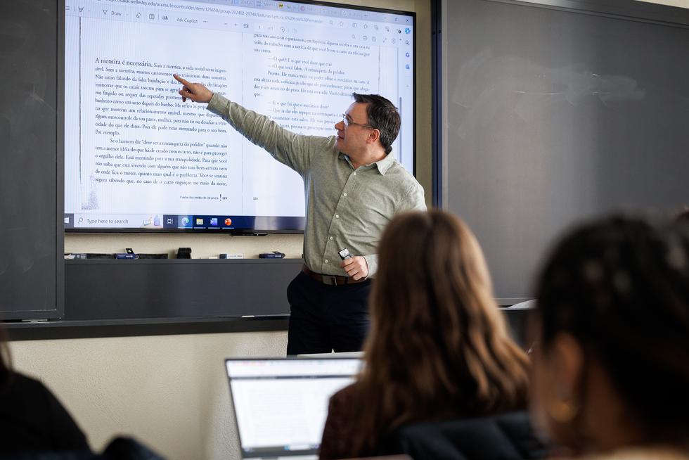 António Igrejas stands at the front of a classroom and gestures at something written on the screen. The screen shows a scan of a book written in Portuguese.