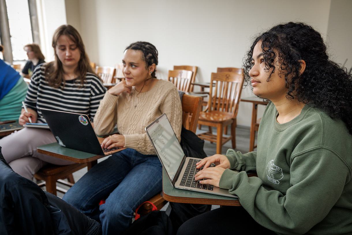 A student looks up from her desk thoughtfully while two students in the background confer on notes.