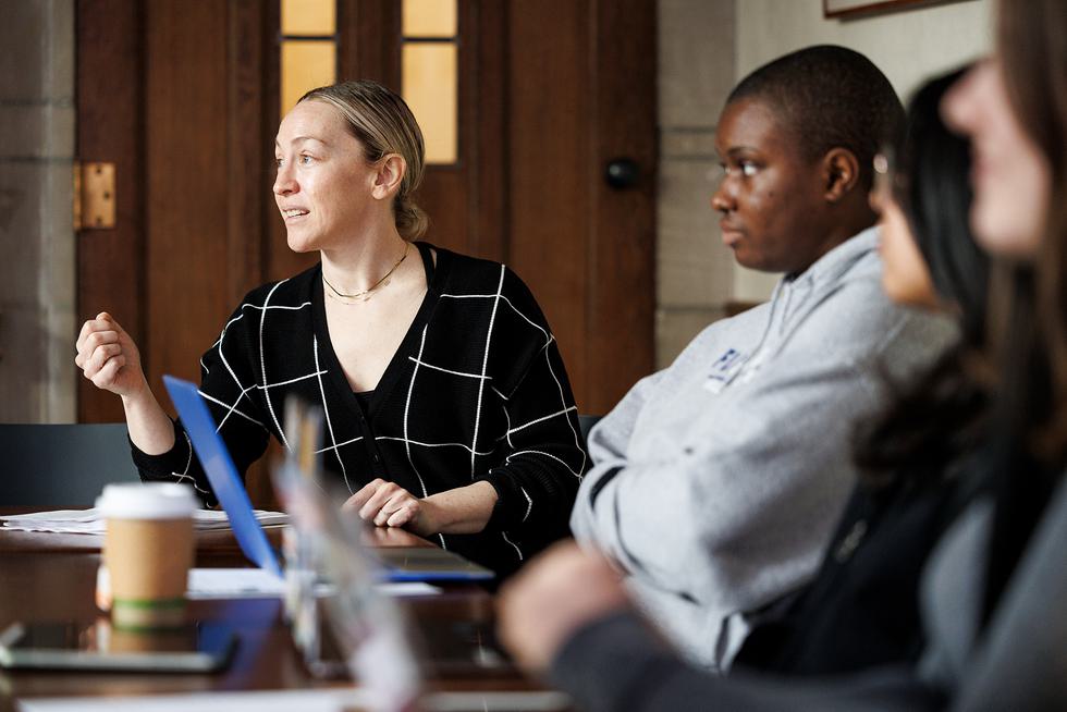 Jennifer Musto sits at a table with students and speaks to them. She is wearing a black and white striped cardigan.