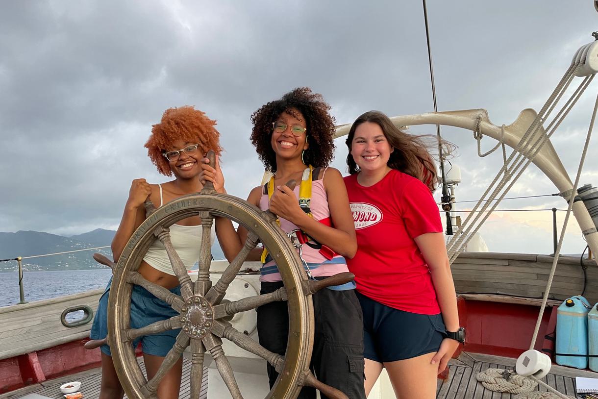 Three students pose, smiling at the helm of a ship.