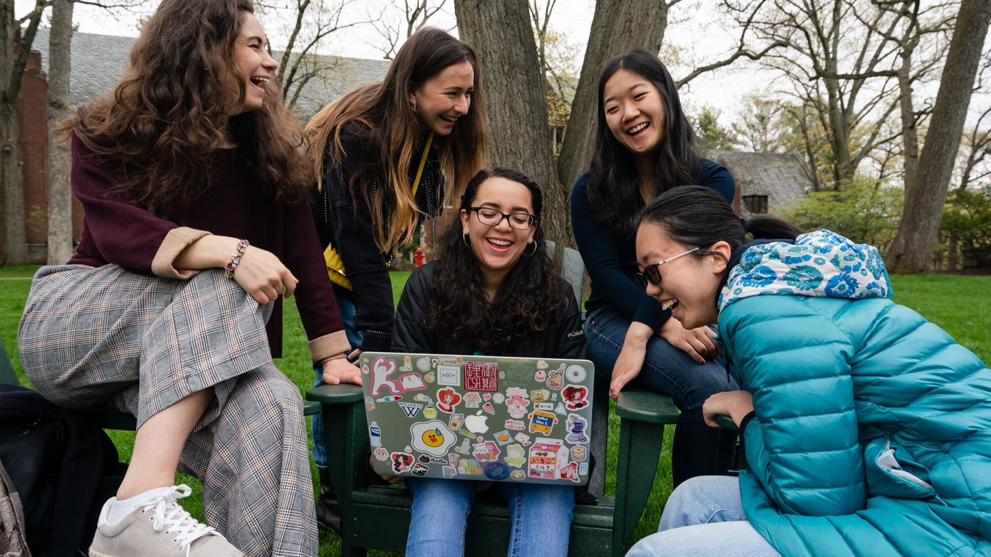 A group of students laugh while watching something on a laptop outside.