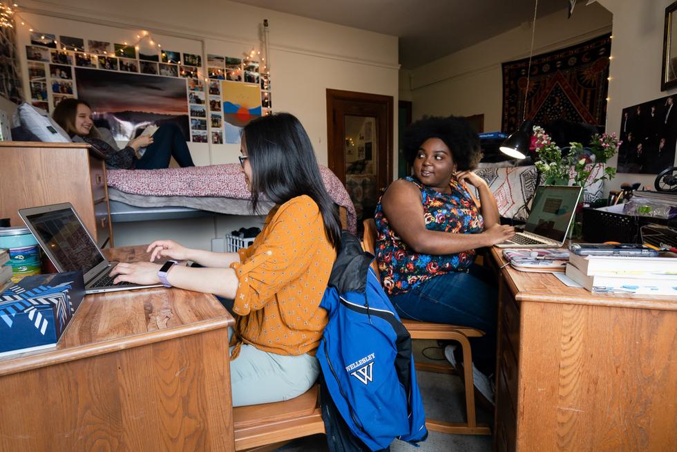 Students in a dorm room work on their laptops at their desk as another student reads in bed.