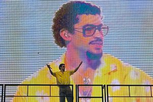Bad Bunny gestures with his hands and stand in front of a jumbotron image of him from the concert he’s perfroming.
