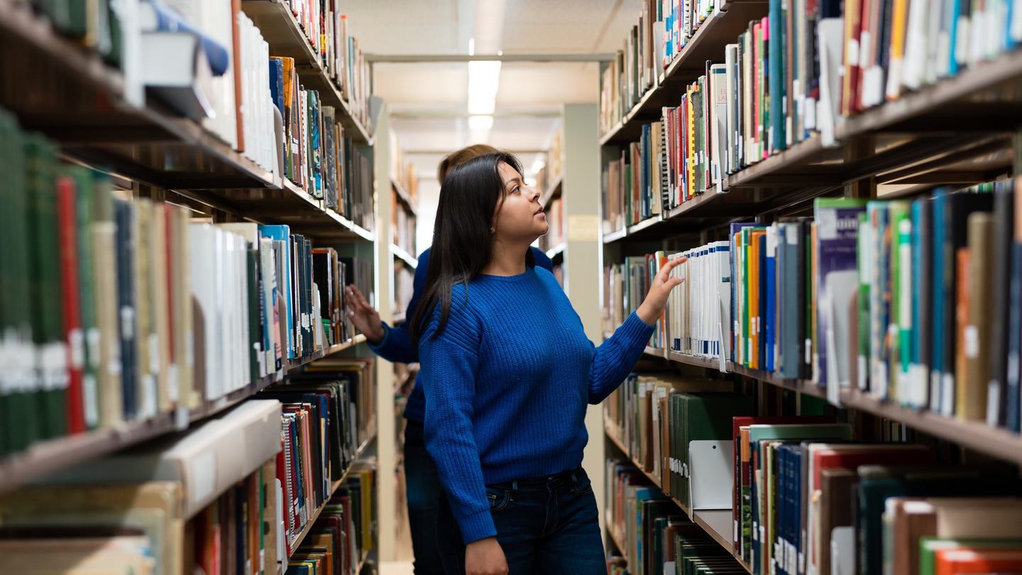 Alt: Students walk between the library's bookshelves, looking at books.